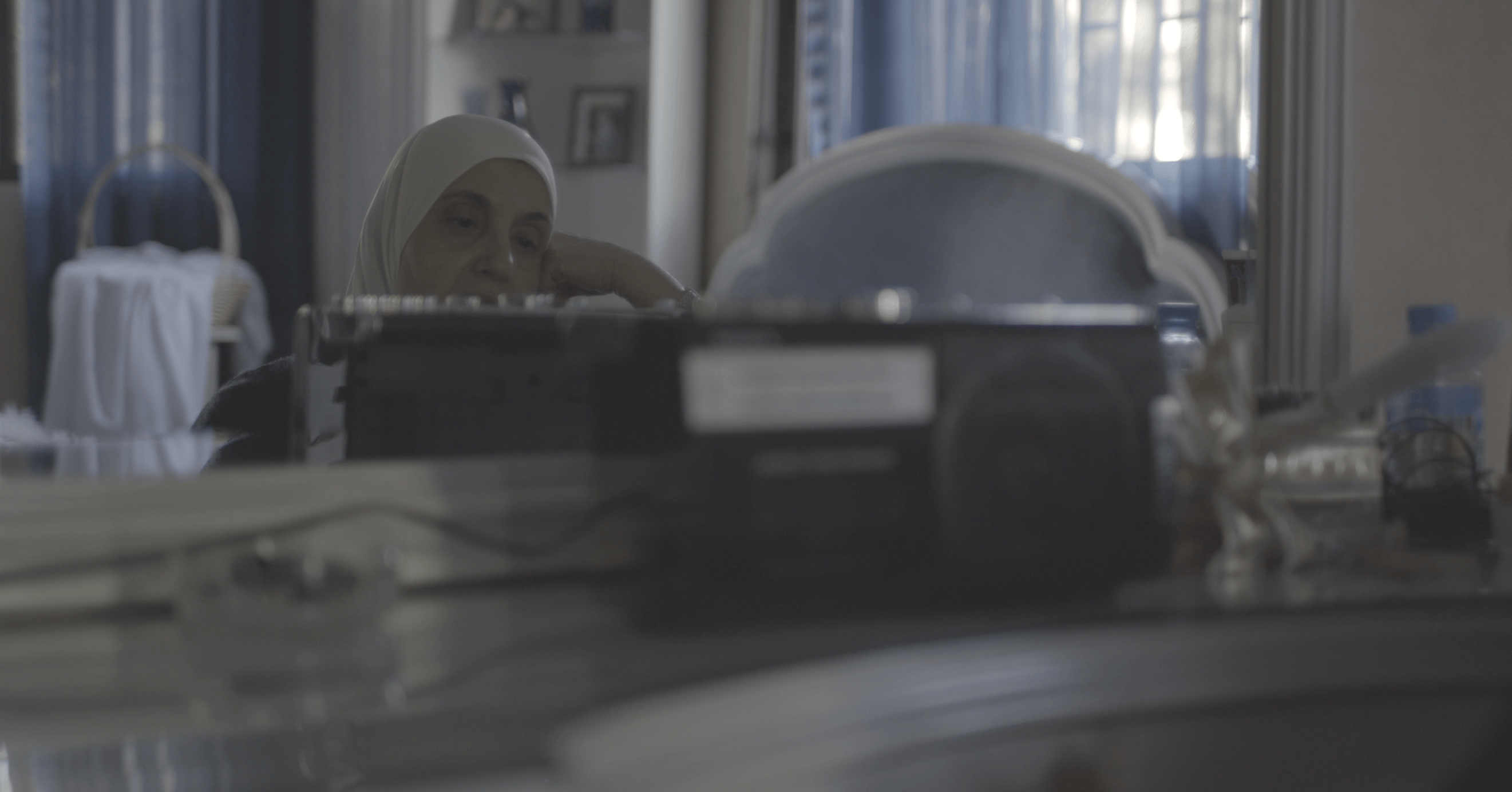 Still from Q. The reflection of a woman wearing a hijab sitting at a desk inside a bedroom with her hand on her face. The reflection of her face is blocked by a small machine sitting on the counter.
