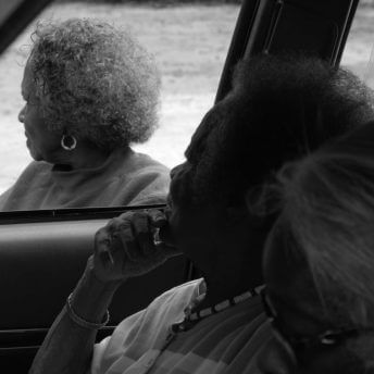 Still from Seeds. Two elderly women are sitting inside of a car, another elderly woman is outside the window. All of them look away from the camera. Black and white shot.