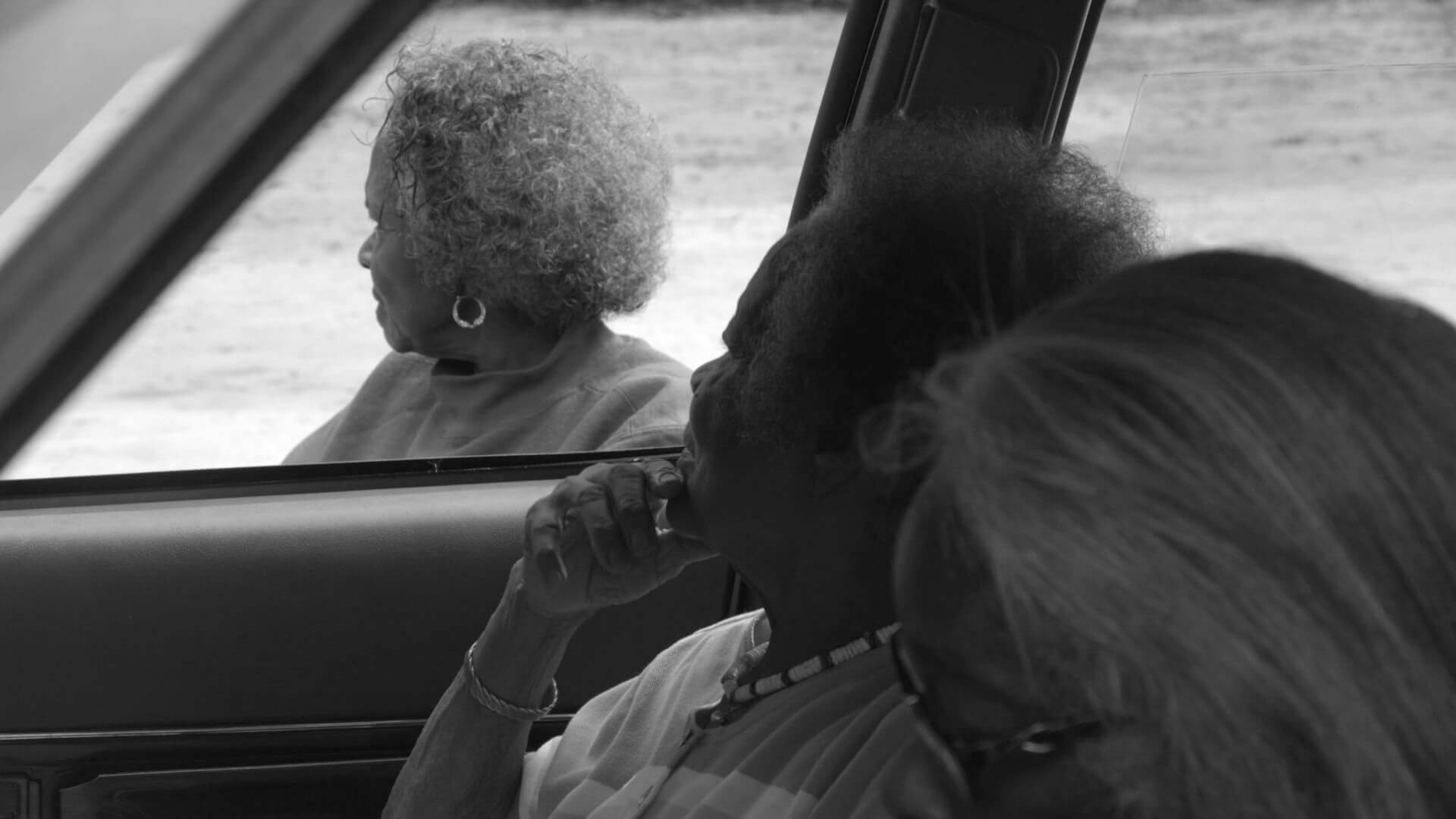 Still from Seeds. Two elderly women are sitting inside of a car, another elderly woman is outside the window. All of them look away from the camera. Black and white shot.