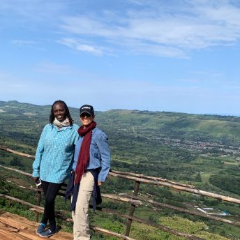 Zippy Kimundu and Meena Nanji stand side by side looking directly into the camera. They stand on a wooden platform in front of a wooden fence. Below them is a faraway landscape of a green valley.