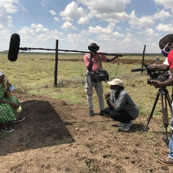 One gaffer and one DP stand on either side of Zippy Kimundu in the middle of an open plain. All three face a woman to the left left who is seated and wearing a long green dress.