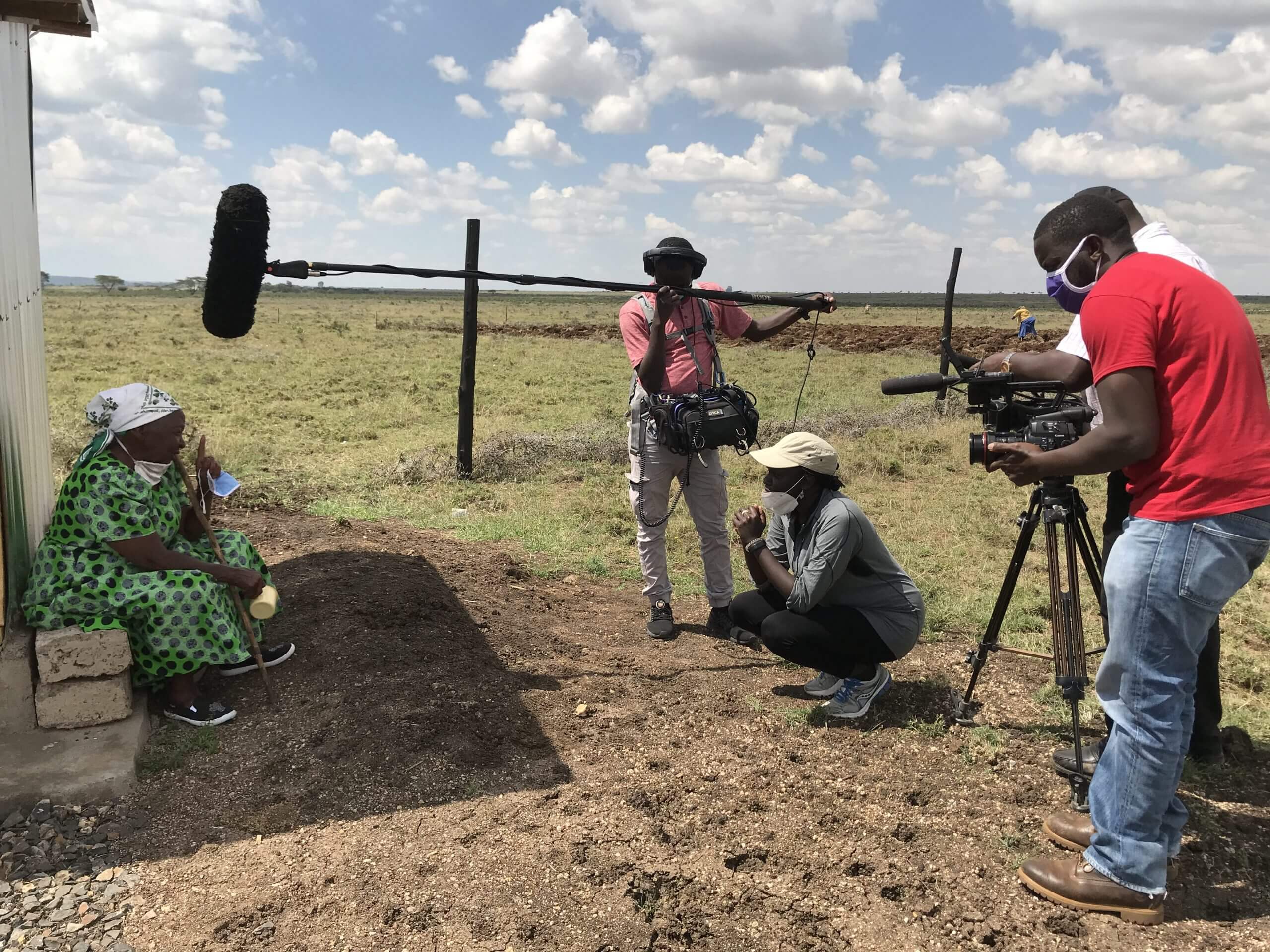 One gaffer and one DP stand on either side of Zippy Kimundu in the middle of an open plain. All three face a woman to the left left who is seated and wearing a long green dress.