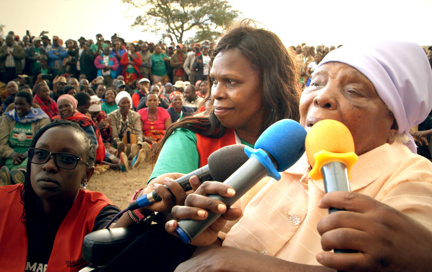 Still from Testament. An older woman speaks into three microphones being held to her mouth. Two other women, stand to the side of her, facing the camera. In the background, many people are sitting in a field looking at the three women.