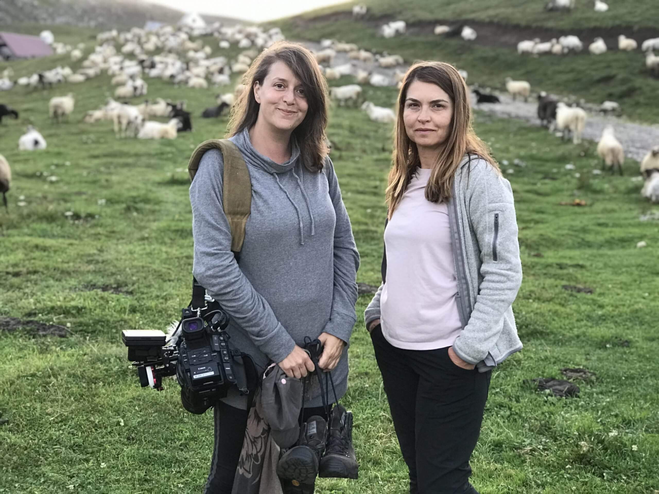 A production still from The Last Nomads. Two people stand in a field looking directly at the camera. Behind them are a patch of green grass and many sheep roaming around. One of them has a large digital camera lung over their shoulder and is holding additional camera equipment.
