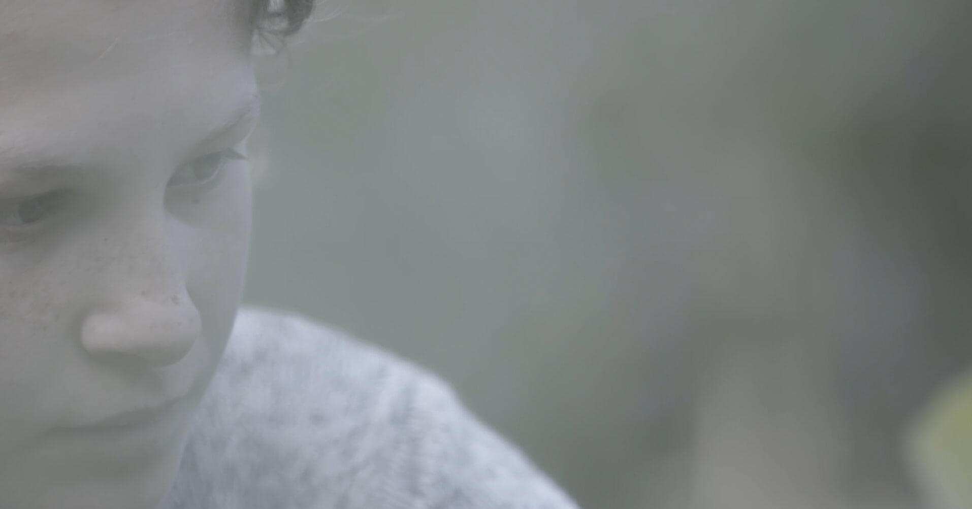 A still from The Last Nomads. Close-up shot of a person's face. They are looking down slightly, although still facing the camera. The image is hazy and slightly out of focus. The person is situated in front of a green background.