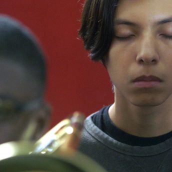 Still from The Tuba Thieves. Close-up of a person with eyes closed in front of a red background. Beside them is a Tuba player out of focus. Color photograph.