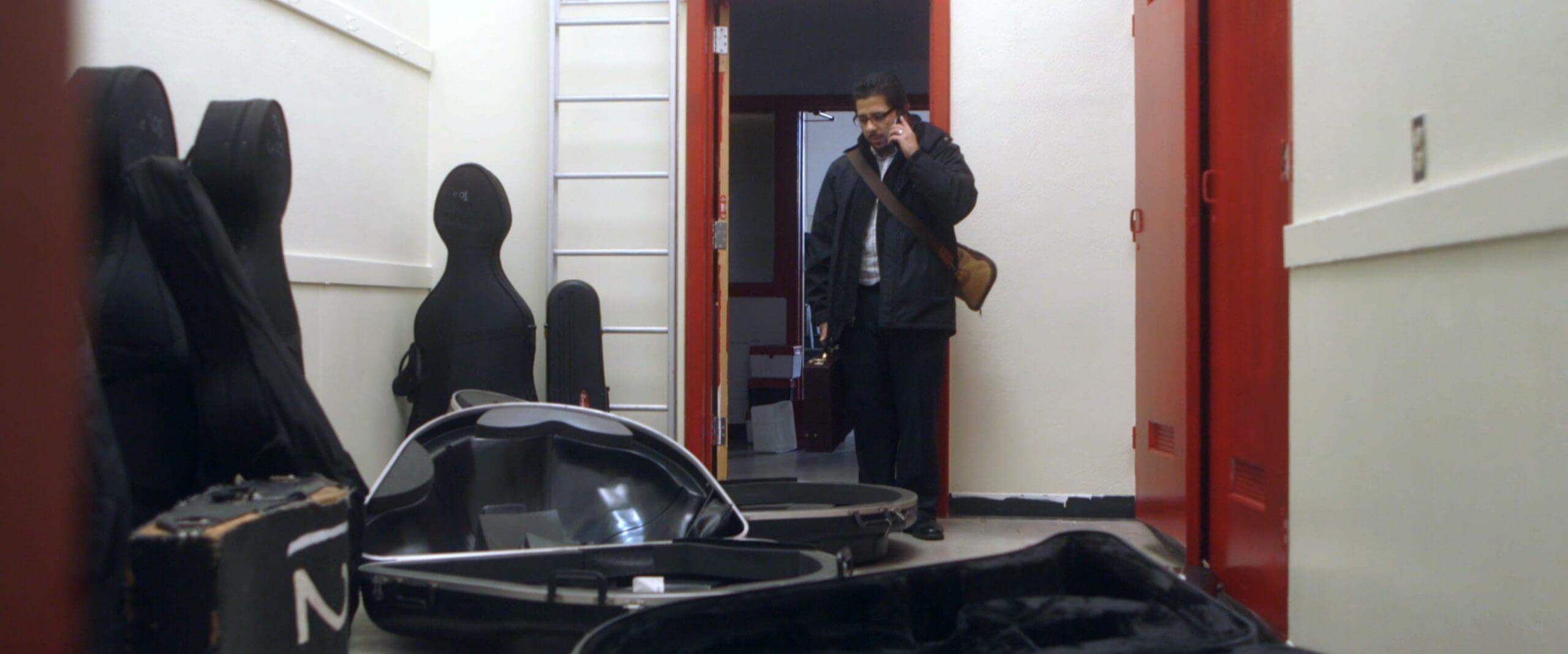 Still from The Tuba Thieves. A person in a jacket on a phone call standing in the doorway of a room. The room is filled with empty instrument cases. Color photograph.
