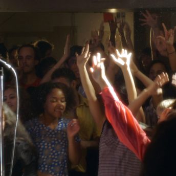 Still from The Tuba Thieves. Group of people with their hands raised. Color photograph.