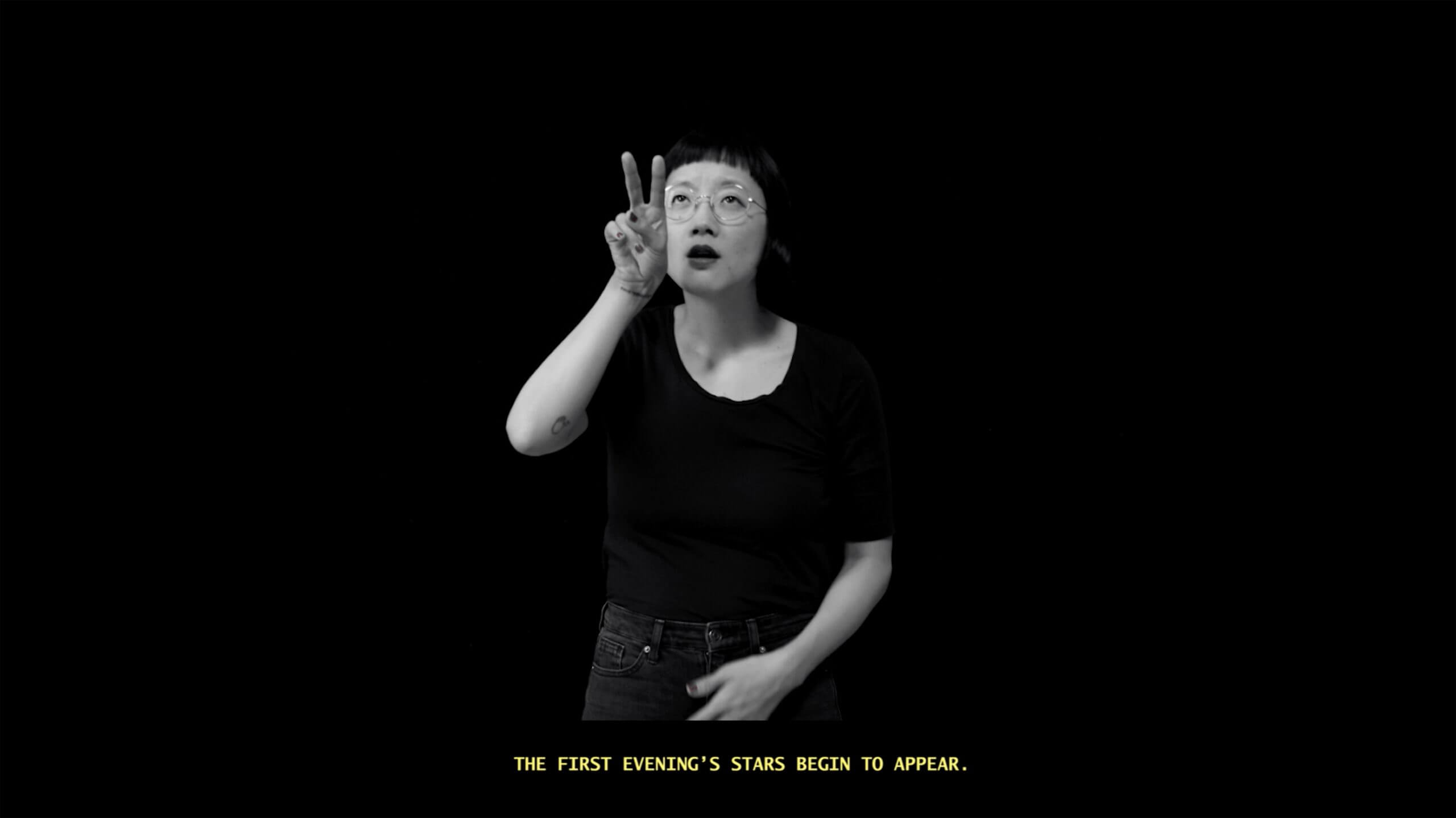 Still from The Tuba Thieves. A person looking up with their hand beside their face holding two fingers. Below them is a text that reads the first evening's stars begin to appear. Black and White photograph.