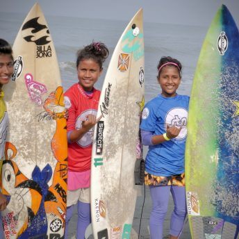 Still from Bangla Surf Girls. Three young girls are wearing aquatic clothing and holding a surf board. They smile at the camera.