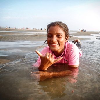 Still from Bangla Surf Girls. A young girl is lying on the seashore, she is smiling at the camera and is partly being covered by the ocean.