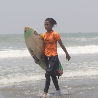 Still from Bangla Surf Girls. A young girl is walking on the shore of the ocean, she is holding a surf board with one arm.
