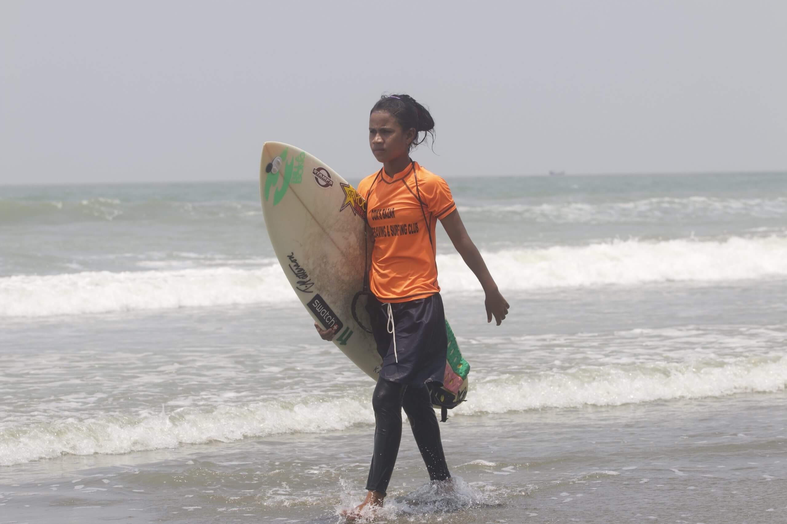 Still from Bangla Surf Girls. A young girl is walking on the shore of the ocean, she is holding a surf board with one arm.