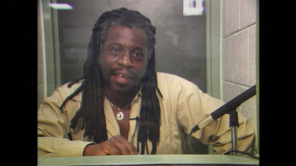 Archive photograph of Dr. Mutulu Shakur. He is talking from behind a glass screen.