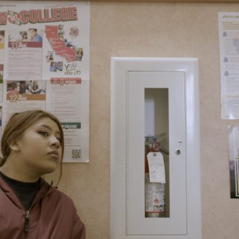 Still from Fruits of Labor. Film protagonist, Ashley, a young woman, is looking in an off-screen direction and is wearing a burgundy jacket and standing against a beige wall that has taped posters on which text can be read "How to get to college" and "NYU Precollege."