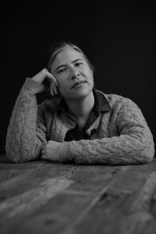 Emily Cohen Ibanez looking straight ahead. She is wearing a thick cardigan sweater, and is leaning her arms on a wooden table, and her head is supported on her right hand. Black and white portrait with black background.