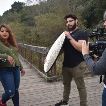 Production still from Fruits of Labor. Film protagonist, Ashley, is leaning with her arm against a wooden walkway. A man to her right holds a reflector directing light towards her. The film director, Emily Cohen Ibanez, is filming with her camera strapped to her chest via a harness.