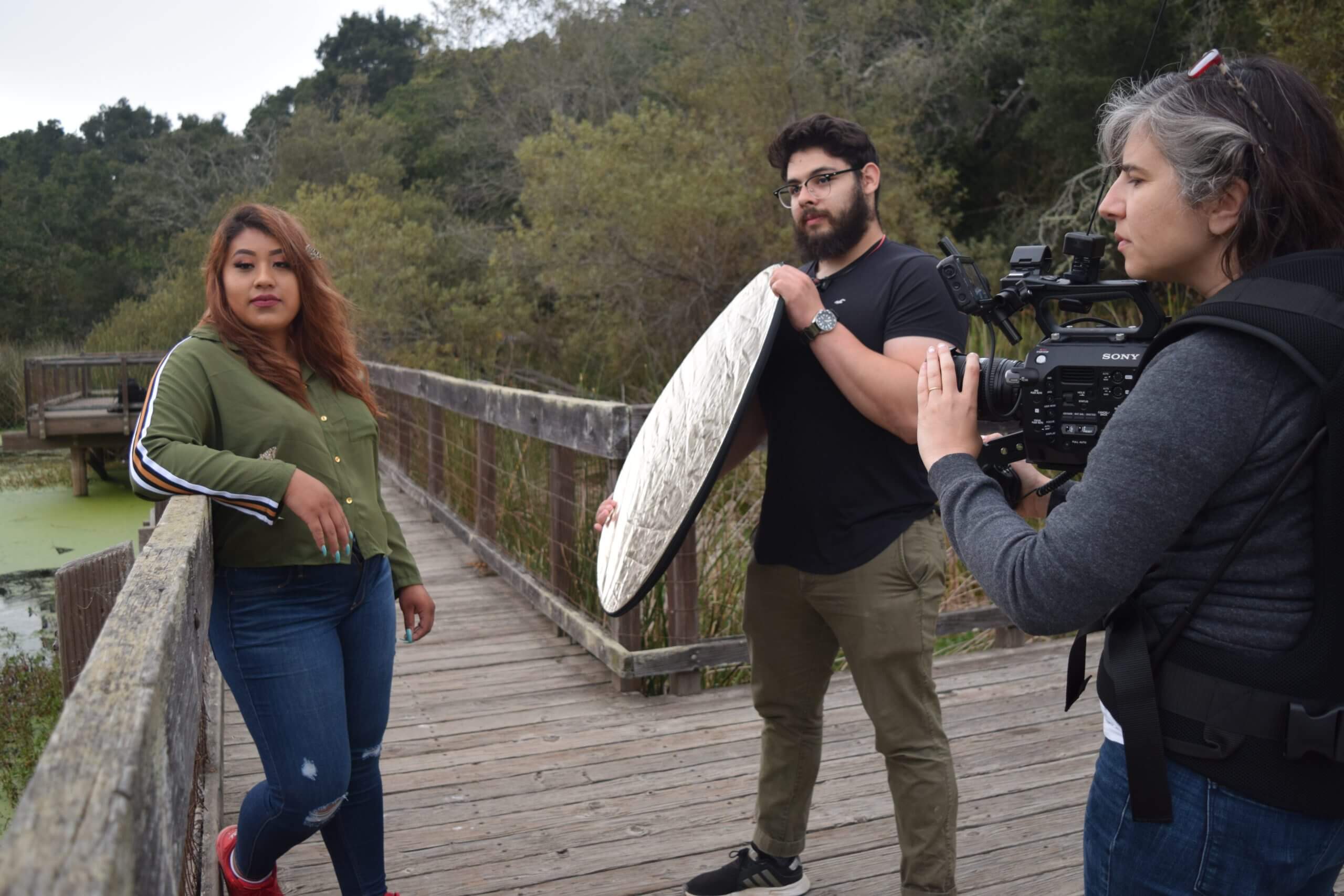 Production still from Fruits of Labor. Film protagonist, Ashley, is leaning with her arm against a wooden walkway. A man to her right holds a reflector directing light towards her. The film director, Emily Cohen Ibanez, is filming with her camera strapped to her chest via a harness.