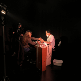 Production still from Fruits of Labor. Black film set with single light source on the film protagnist, Ashley, who is sitting at a table and haivng her nails filed and painted by a nail technician. The film director, Emily Cohen Ibanez, is filming from behind the tripod camera.