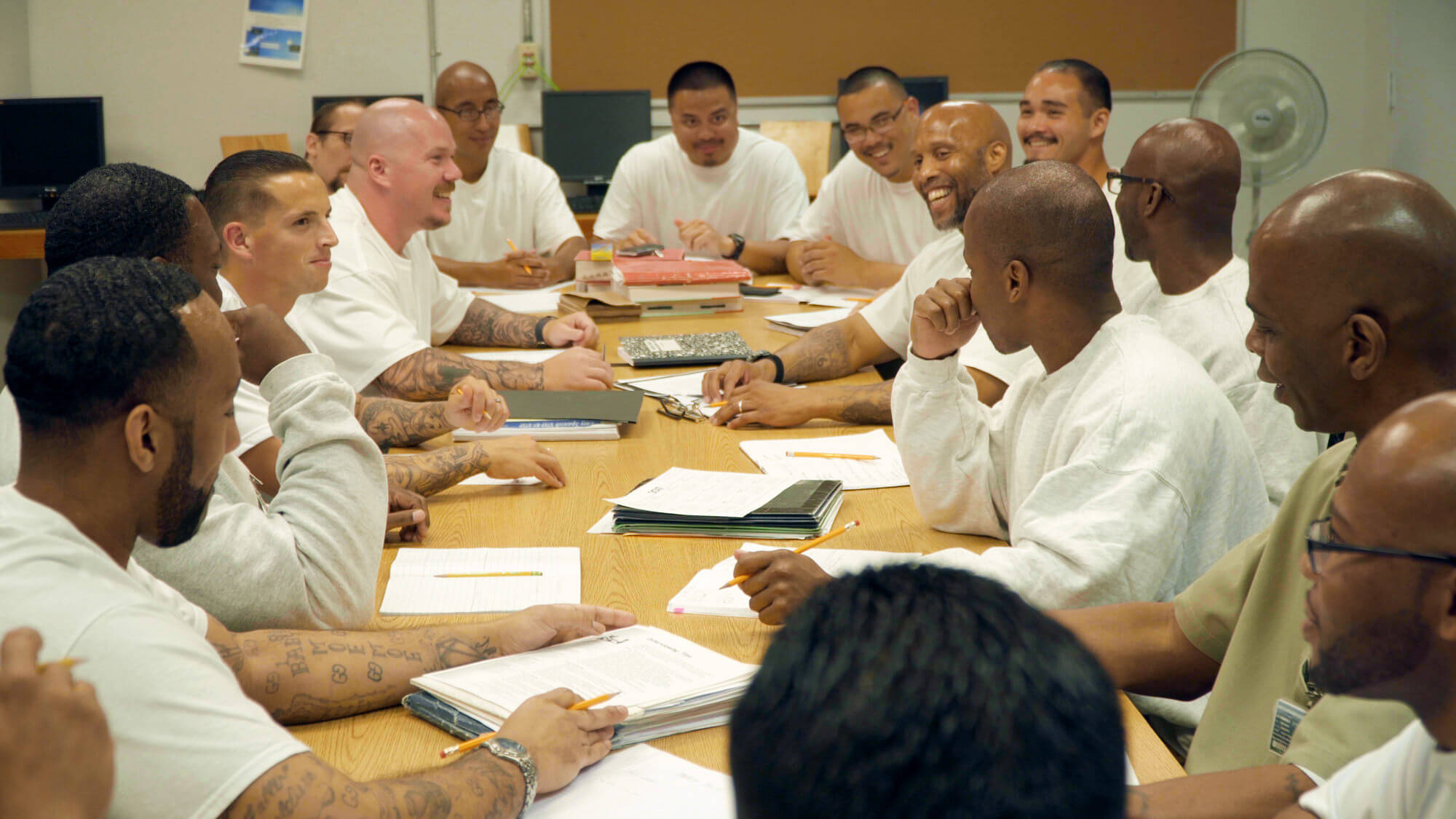 Still from Since I Been Down. A group of men in prison is sitting at the table, they have papers and pencils. Some of them smile.