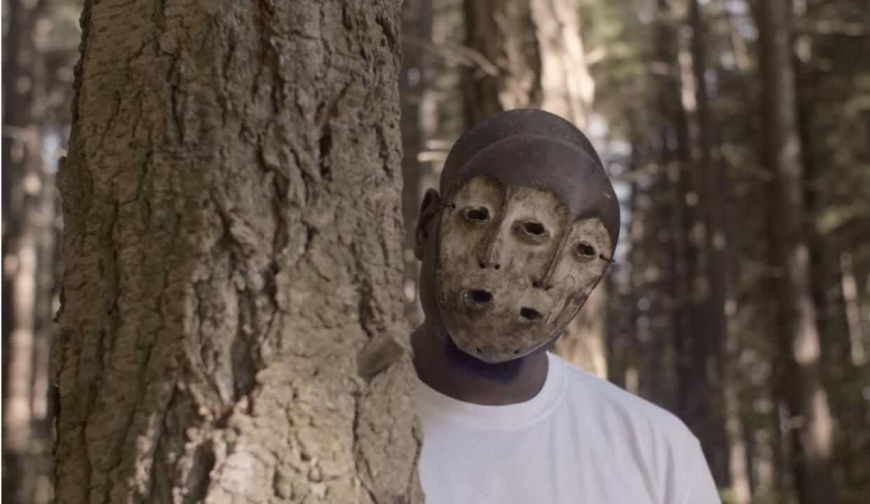 Still from Since I Been Down. A person wearing a mask peeks behind a tree.