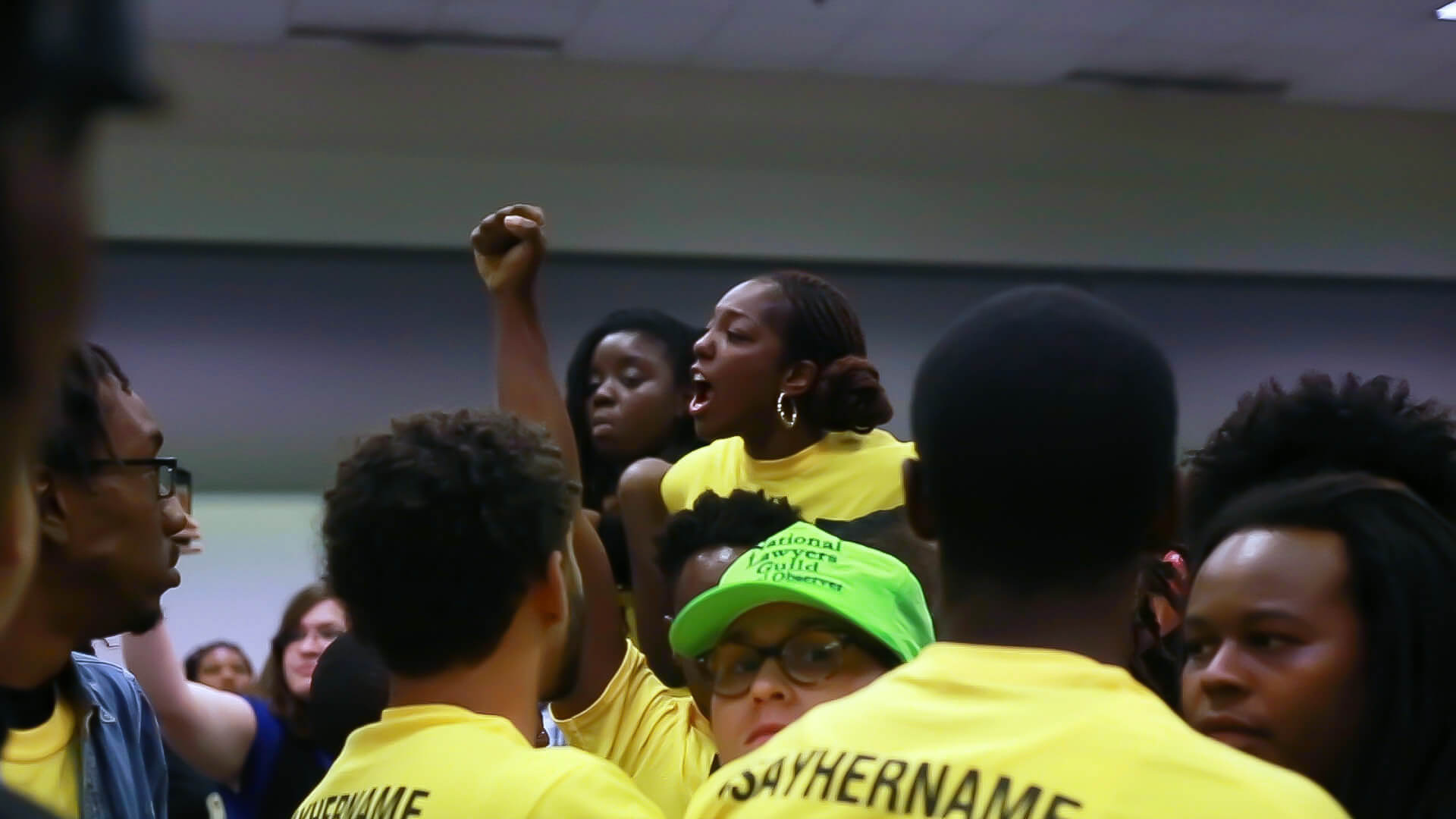 Still from Unapologetic. People gathered, wearing yellow shirts, and one of them has their fist up in the air. A woman stands out from the crowd.
