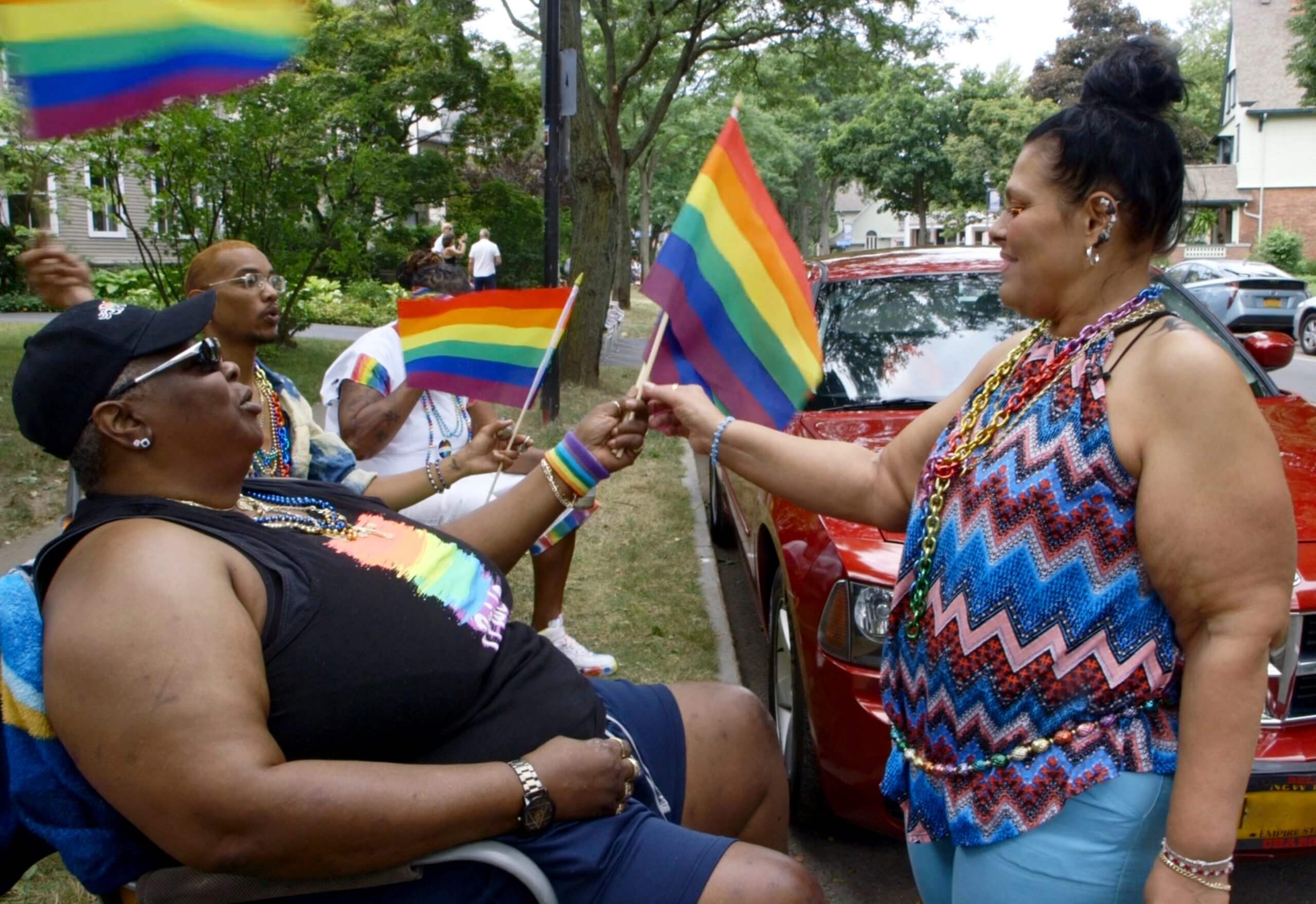 Still from And So I Stayed. Three people are sitting on chairs, holding LGBTQ+ flags, and a woman is standing in front of one of them and grabbing one of the flags. There is a red truck parked.