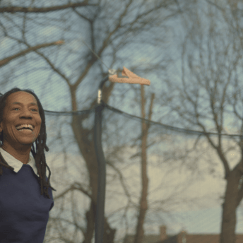 Still from I'm Free Now You Are Free. A woman with chest-length dreads and wearing a navy-blue sweater with a white-collared shirt, is smiling widely. She is seen through the mesh of a trampoline net.
