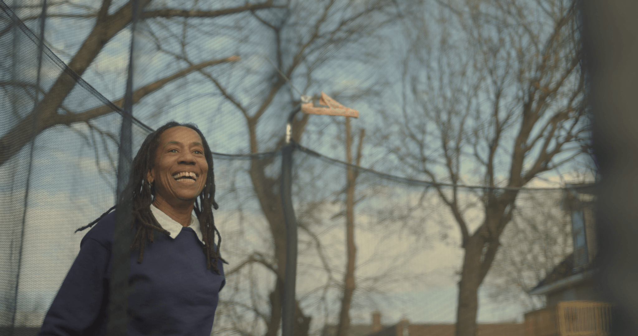 Still from I'm Free Now You Are Free. A woman with chest-length dreads and wearing a navy-blue sweater with a white-collared shirt, is smiling widely. She is seen through the mesh of a trampoline net.