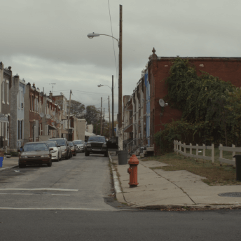 Still from I'm Free Now You Are Free. A wide shot of a city street. Two-story buildings line both sides of the street, and a fenced-in green area is to the right. The sky is overcast, and a man is walking on side of the street.