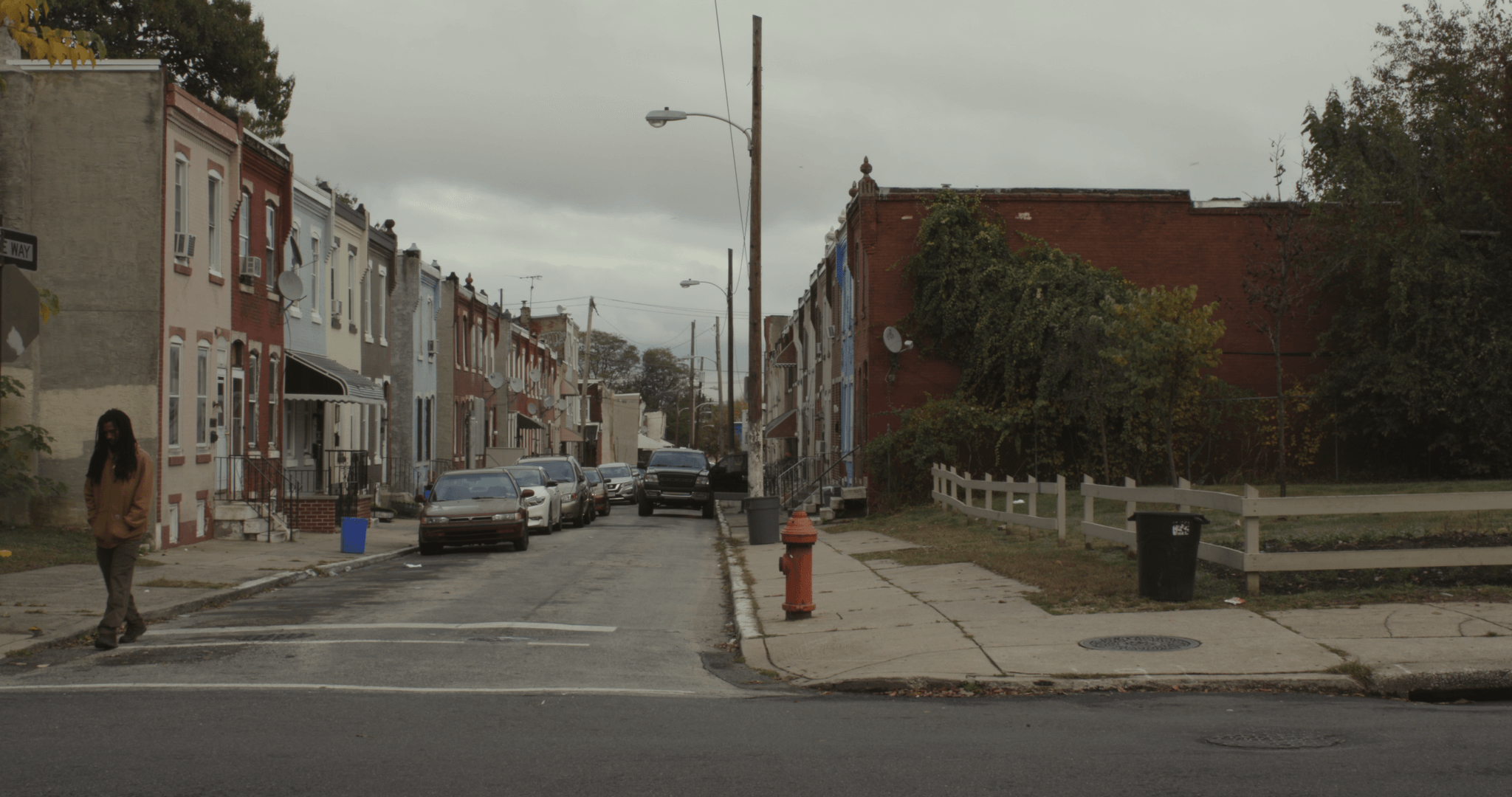 Still from I'm Free Now You Are Free. A wide shot of a city street. Two-story buildings line both sides of the street, and a fenced-in green area is to the right. The sky is overcast, and a man is walking on side of the street.