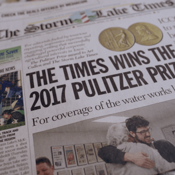 Still from Storm Lake. Close-up shot to a The Storm Lake Times cover, the headliner is "The Times Wins the 2017 Pulitzer Prize for the coverage of the water works lawsuit", the photo on the cover is of two men hugging.