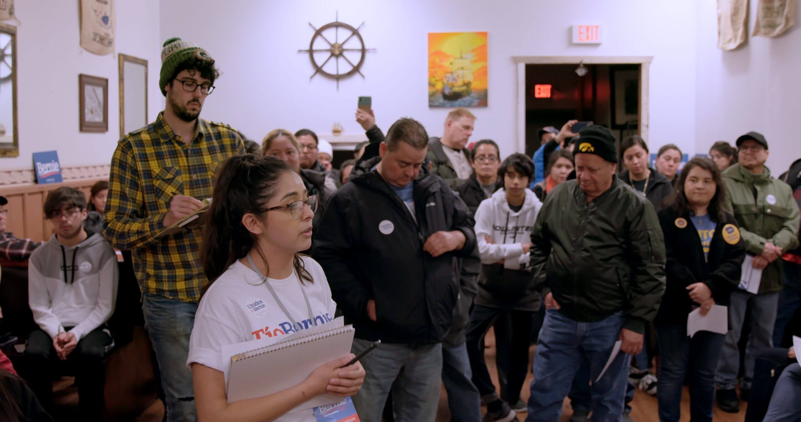Still from Storm Lake. People in a meeting. In the foreground, a young person with a notebook wears a t-shirt that says "Tio Bernie."