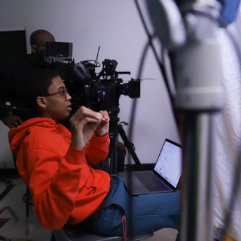 Cai Thomas in a production set. Cai is sitting on a chair, she has a computer on her lap, she is giving instructions, and is moving her hands. Behind her, there is a man with video equipment.