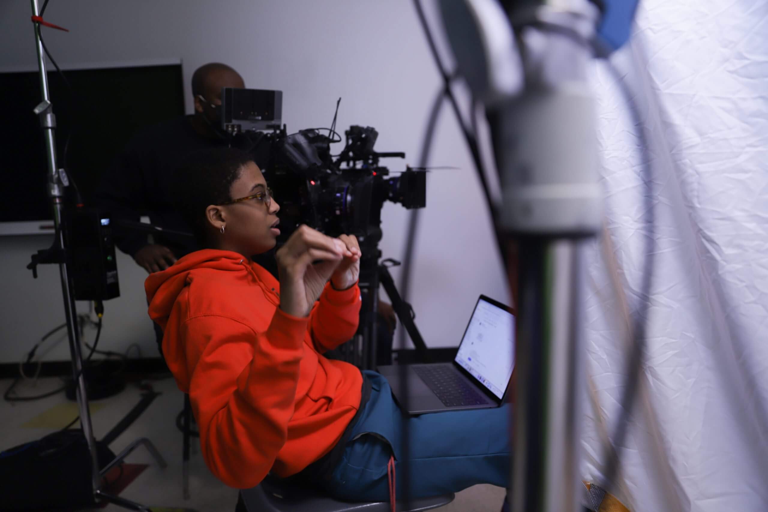 Cai Thomas in a production set. Cai is sitting on a chair, she has a computer on her lap, she is giving instructions, and is moving her hands. Behind her, there is a man with video equipment.