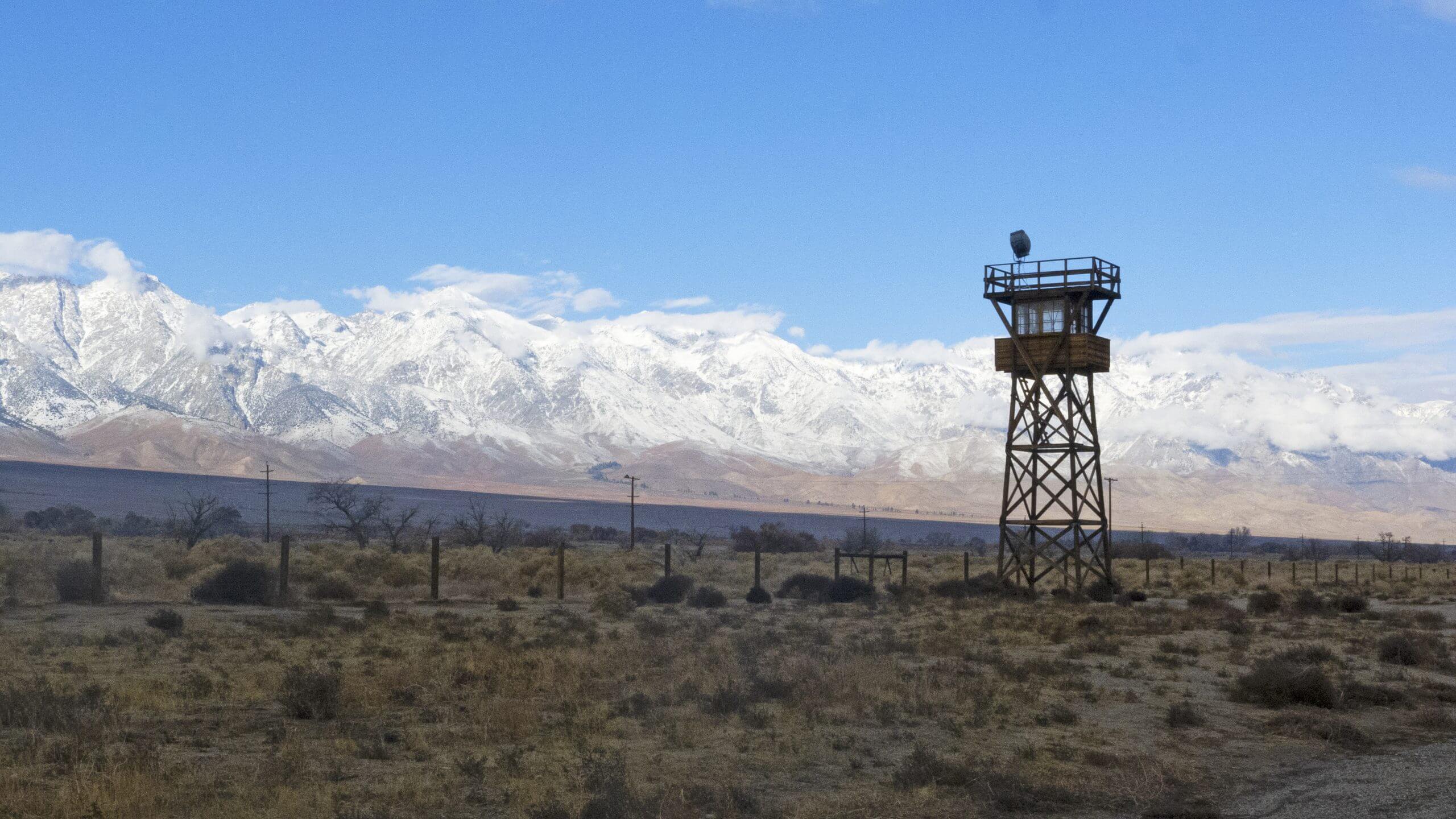 Still from Manzanar Diverted: When Water Becomes Dust. A watchtower stands in a field with a mountain range and blue skies in the background.