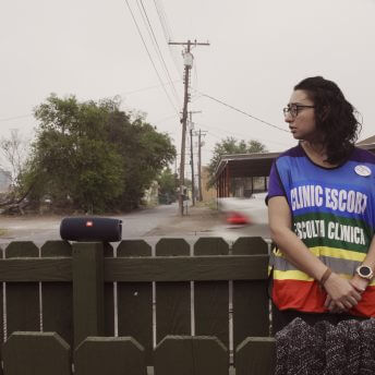Still from On The Divide. A person wearing glasses and a "Clinic Escort" vest leans against a wooden fence next to a portable speaker.