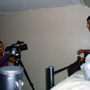 A woman with dark skin and dark hair in a ponytail points a video camera, that's sitting on a tripod, on to a teenager who's sitting on a bunkbed at a shelter.