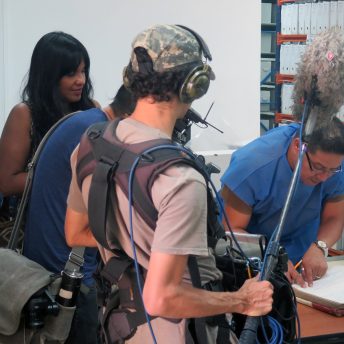 A man with glasses and in a blue uniform leans into a large book that's on a desk while surrounded by a crew of (camera man and sound man) filming him flipping through the book. A woman with dark skin and dark hair stands next to the crew and behind the man they are filming giving directions.