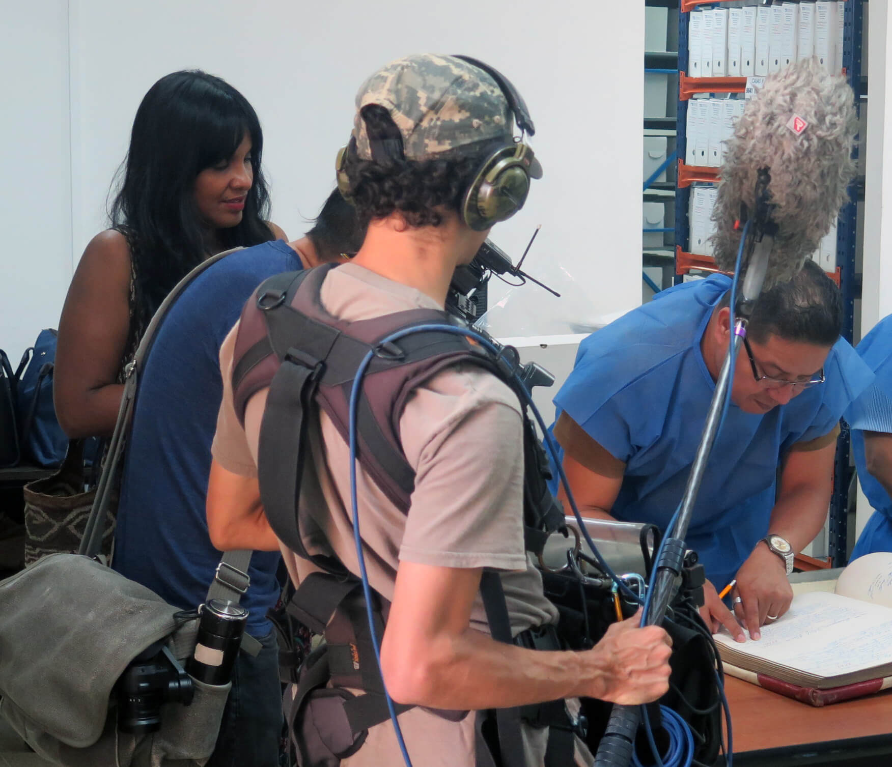 A man with glasses and in a blue uniform leans into a large book that's on a desk while surrounded by a crew of (camera man and sound man) filming him flipping through the book. A woman with dark skin and dark hair stands next to the crew and behind the man they are filming giving directions.