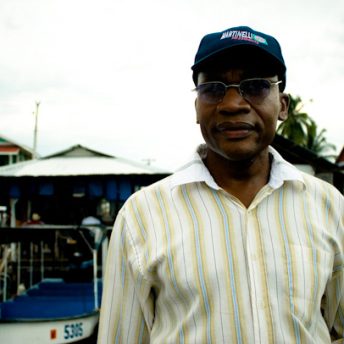 Black man with dark glasses, blue cap and light yellow shirt, stands outdoors looking into the camera, behind him a dock with small boats.