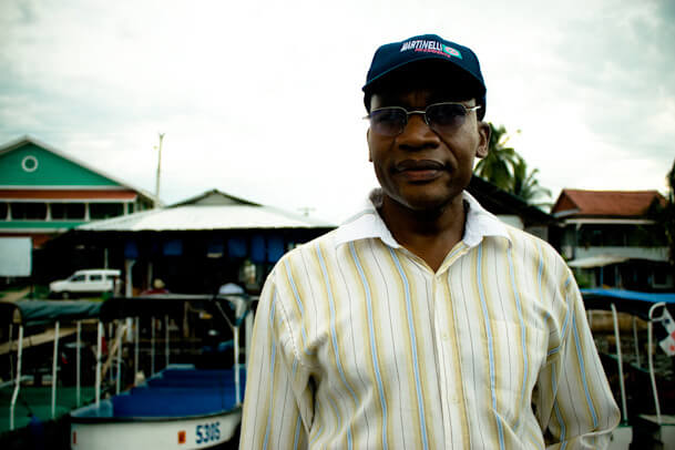 Black man with dark glasses, blue cap and light yellow shirt, stands outdoors looking into the camera, behind him a dock with small boats.