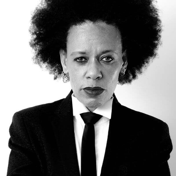 Black & White medium image of Dr Bev in an Afro hairstyle, wearing a black tux and black tie, looking directly at camera.