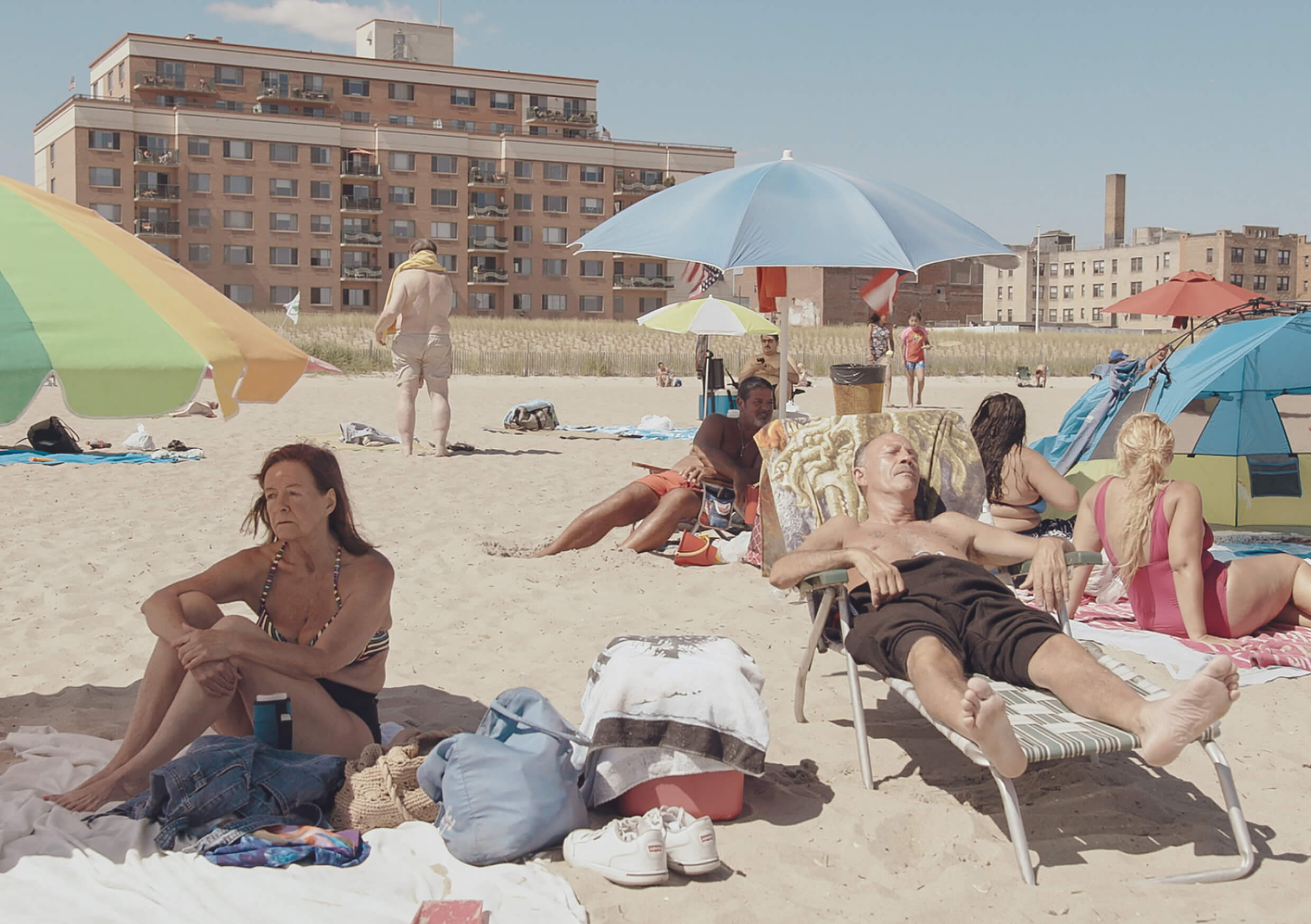 A older, white couple sit and lie down on the beach in a crowd of sunbathers and umbrellas.