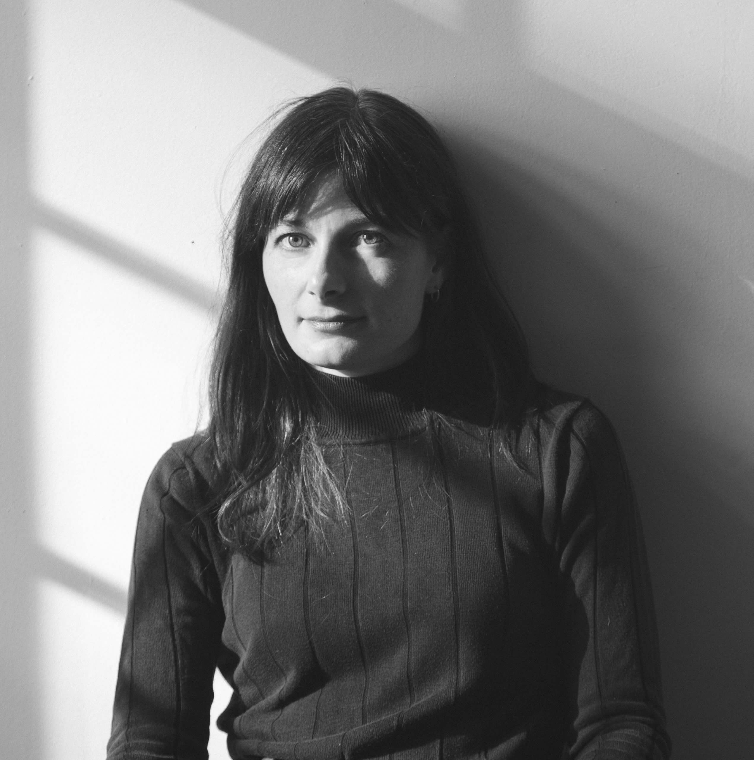 This is a headshot of Brett Story, a white woman with freckles and long brown hair and bangs. She is sitting at a table against a white wall, wearing a black turtleneck and jeans.