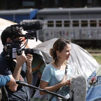 Petra Costa and João Atala in the middle of a shooting. Atala is holding a camera, Petra is facing forward and and we can see tents, a train in the background and a hand in the corner holding the boom.