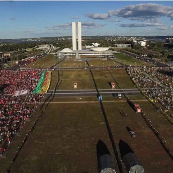 Drone shot of Brasilia and the building that houses both the Senate and Chamber House of Deputies, consisting of what we consider the Congress in Brazil. On one side there is a crowd mostly dressed in red representing the left and on the other side a crowd mostly dressed in green and yellow representing the right.