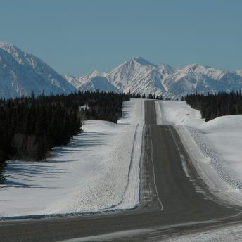 Highway lined with snow and pine trees extends toward snow-covered mountains in the distance.