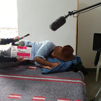 A woman holds a clapperboard while a man sleeps on a bed, hiding his face with a hat