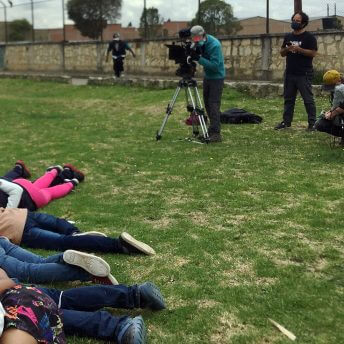 A production crew works in an open field, while a group of young women are laying on the grass